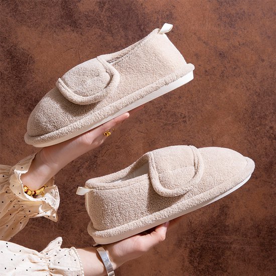 Handen houden twee beige pluche pantoffels met verstelbare klittenbandsluiting vast.