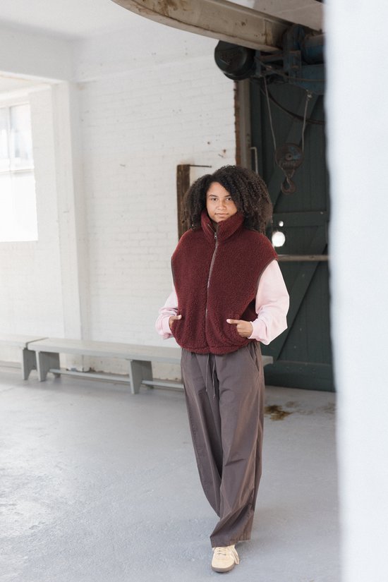 Model draagt een bordeauxrode wollen bodywarmer over een roze trui en donkere broek.