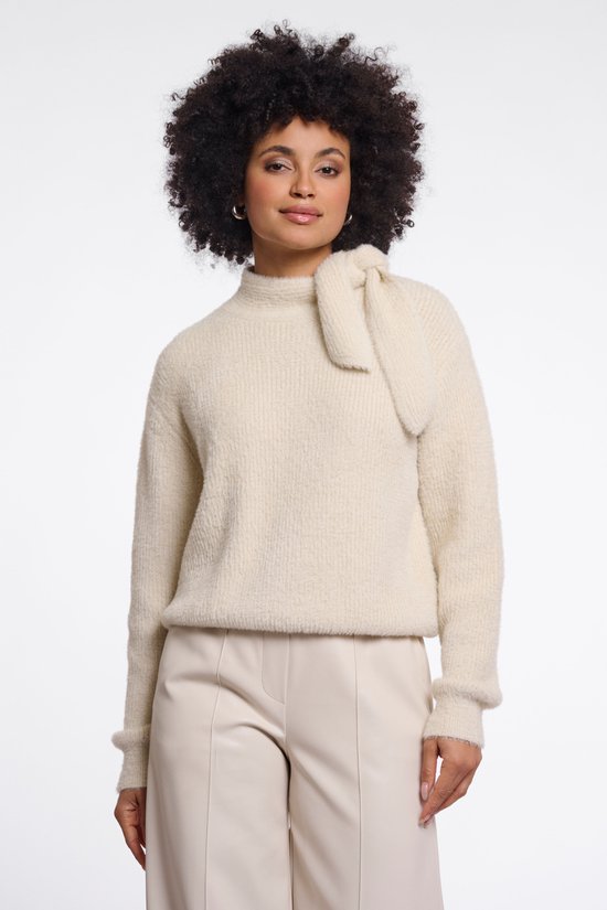 Model draagt een beige sweater met textuur en een knoopdetail bij de hals, gecombineerd met een lichte broek.