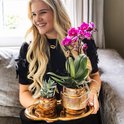Een glimlachende vrouw houdt een gouden dienblad vast met een paarse orchidee en een vetplant in gouden potten.