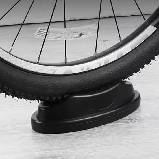 Zwarte voorwielsteun onder een fietswiel, met een plat profiel en verhoogde zijkanten. Het product is geplaatst op een houten vloer.