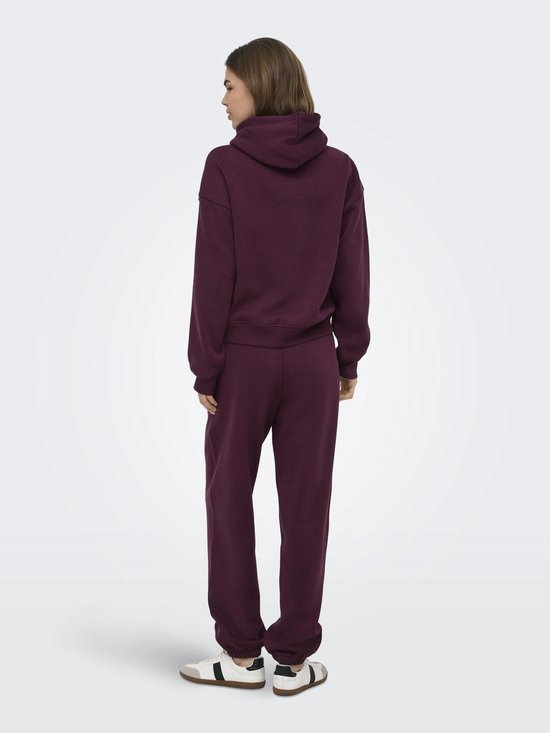 Vrouw staat met de rug naar de camera, draagt een bordeauxrode hoodie en bijpassende joggingbroek.