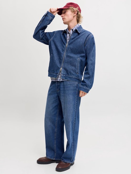 Man met korte blonde haren, donkerrode pet, jeansjack en wijde jeans, hand op pet.
