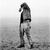 Man op een strand met een denim jas en een spijkerbroek, de hand voor zijn gezicht.