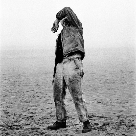 Man op een strand met een denim jas en een spijkerbroek, de hand voor zijn gezicht.