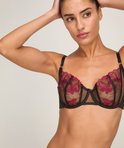 Model in zwarte beha met rode bloemenkant en zwarte bandjes. Ze legt haar handen in haar haar.