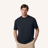 Man met bruin haar, handen in de zakken, in een donkerblauw T-shirt en beige broek.