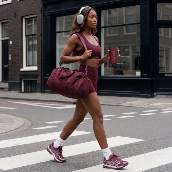 Vrouw met bordeauxrode sportkleding en tas, met hoofdtelefoon en beker, op een zebrapad.