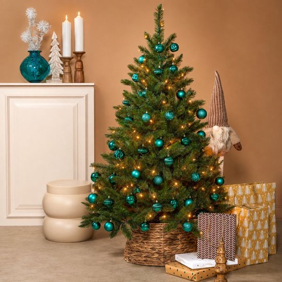 Groene kunstkerstboom versierd met teal kerstballen en lichtjes, naast een open haard.