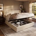 Beige gestoffeerd bed met opengeklapte opbergruimte, vol met manden en opgevouwen spullen. Het bed is opgemaakt met een plaid.