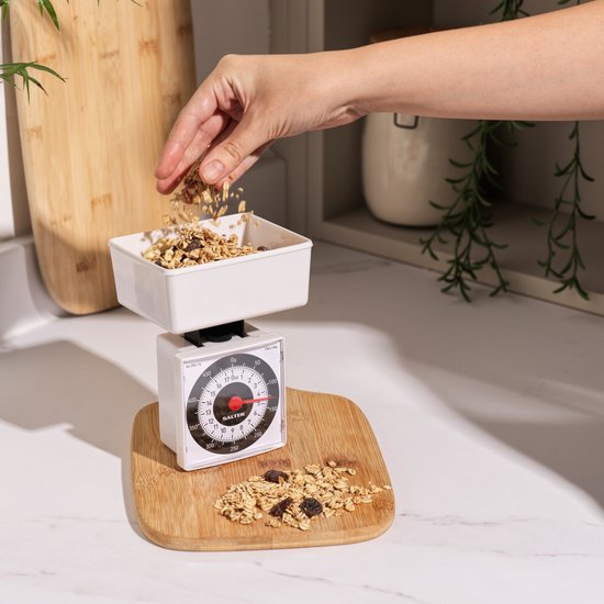 Witte keukenweegschaal met een ronde bak waarin muesli wordt gedaan, met muesli ernaast.