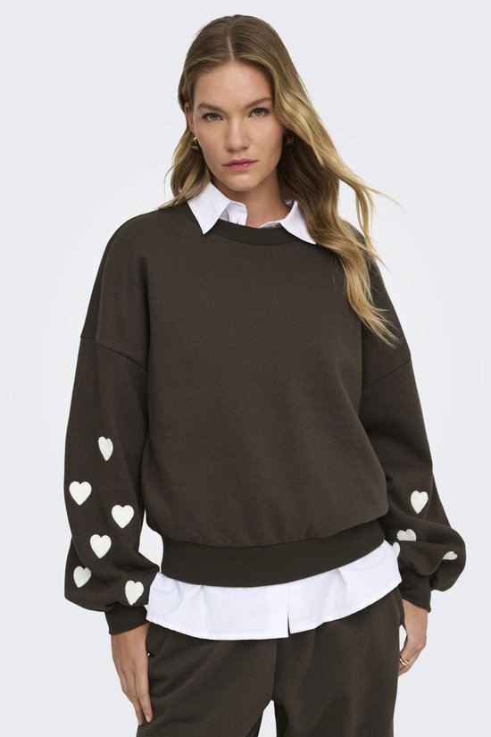Donkerbruine sweater met witte hartjes op de mouwen, gedragen door een model met een witte blouse eronder.