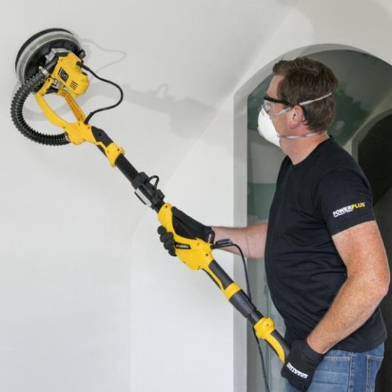 Man met veiligheidsbril en mondkapje gebruikt een gele en zwarte schuurmachine op een plafond.