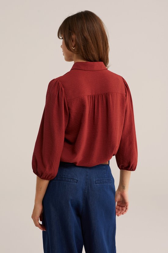 Een vrouw met lang bruin haar, teruggekeerd, draagt een roestbruine blouse met driekwart pofmouwen, een kraagje en knoopjes. Ze draagt een donkerblauwe broek.
