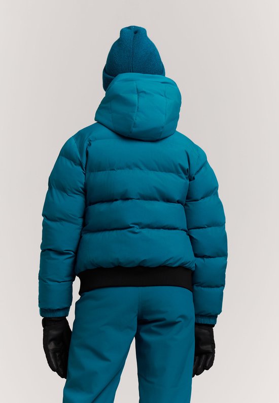 Achterkant van een meisje in blauwe ski-jas, muts, broek en zwarte handschoenen. Zwarte band onderaan de jas.