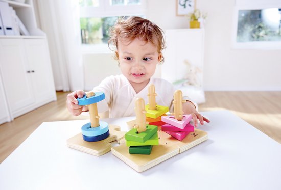 Jong kind met krullend haar speelt met een houten vormpuzzel op tafel. Stapelt blauwe ringen op de peg.