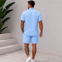 Man, achterkant, draagt lichtblauwe korte mouw blouse en korte broek. Blouse met structuur. Witte sneakers.