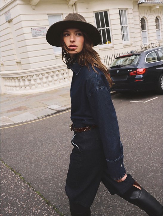 Model draagt bruine vilten fedora met zwarte band en gouden bitdetail.