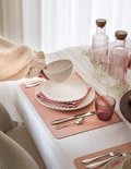 Une main pose un bol blanc sur un set de table rose à côté d'un verre à eau rose.