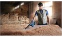 Man in beige vest blaast houtstof weg met blauwe Bosch bladblazer in een werkplaats.