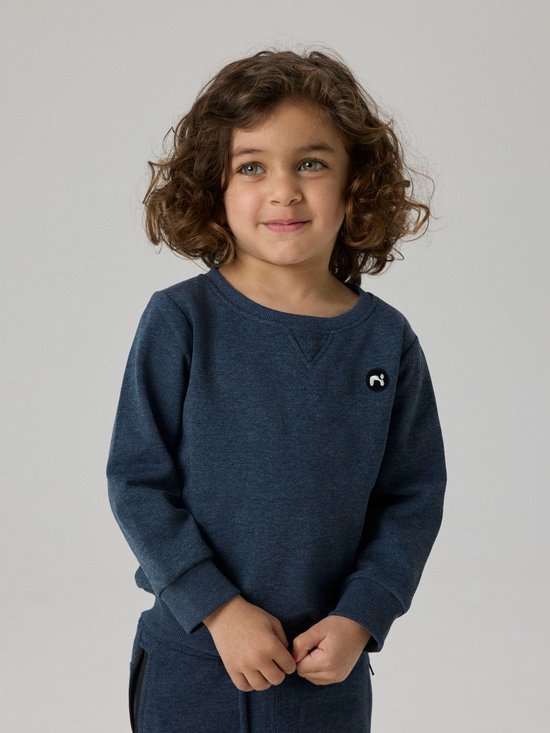 Jongetje met krullend haar, lichtgroene ogen en een donkerblauwe sweater met een geborduurd logo.