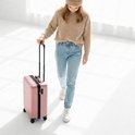 Vrouw met roze koffer op wielen, lopend in een witte ruimte. Ze draagt een beige trui en muts.