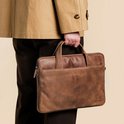 Man met trenchcoat die een bruine leren laptoptas draagt.