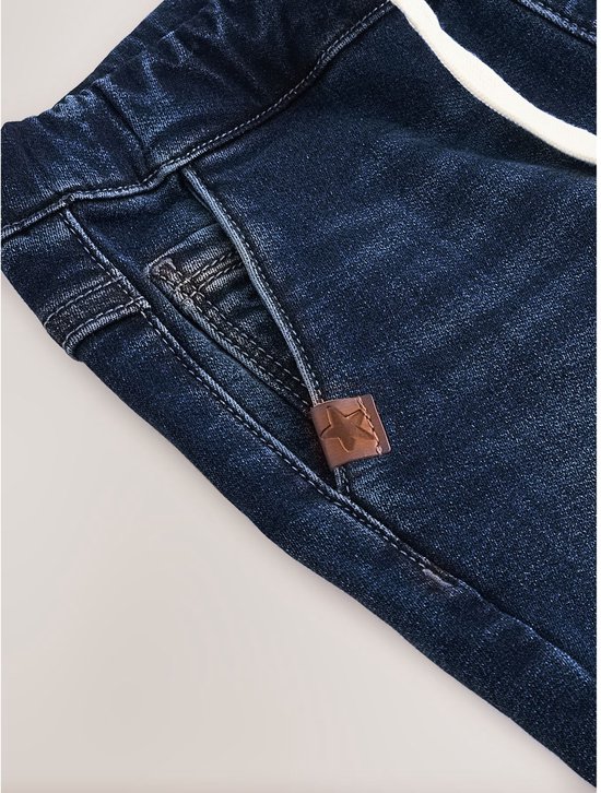 Detail van een donkerblauwe spijkerbroek met trekkoord, zak en decoratieve sterpatch.