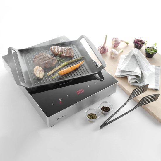 Inductie grillplaat met ribbelgrill, waarop twee steaks, asperges, wortel en knoflook liggen. Naast de grill staan peper- en zoutvaatjes, tang en kruiden.