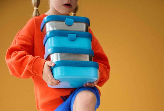 Un enfant portant un pull orange tient des boîtes à lunch Mepal empilées. Les boîtes sont en plastique bleu et en métal.