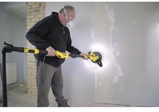 Een man met veiligheidsbril en mondkapje schuurt een muur met een gele en zwarte telescoopschuurmachine.