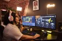 Een streamer met een lichtroze koptelefoon praat in een microfoon aan een bureau met twee monitoren.
