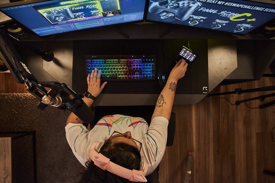 Overzicht van bovenaf van een persoon die aan een bureau zit met meerdere schermen, een toetsenbord, een microfoon en een audiocontroller.