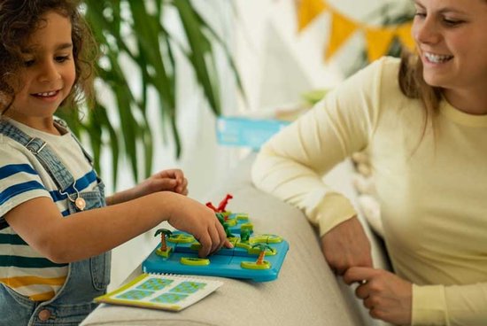 Meisje met krullend haar in spijkerbroek speelt dinosaurusspel met moeder, met rode en groene dino's op een blauw eilandspel.