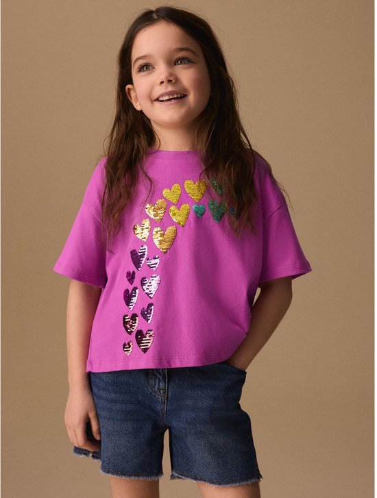 Meisje glimlacht, draagt roze T-shirt met hartjes van pailletten en spijkerbroek.