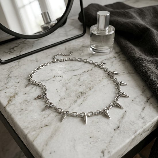 Zilverkleurige schakelketting choker met zeven puntige spikes, op een marmeren oppervlak naast een parfumflesje en spiegel.