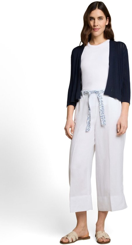 Model draagt een witte top, een witte wijde driekwartbroek met blauwe bloemenriem en een marineblauwe vest.