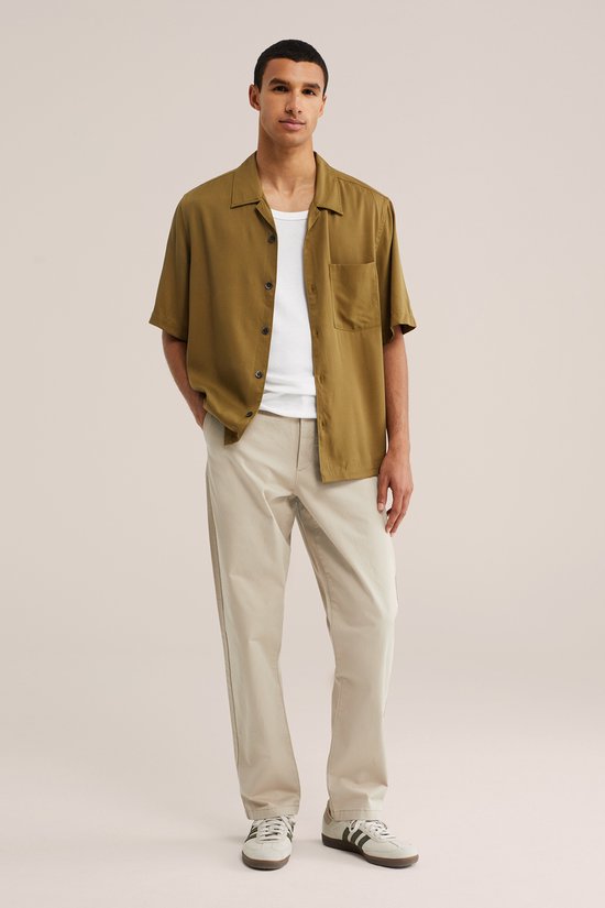 Model staat in een beige chino broek, wit T-shirt en olijfgroen hemd.