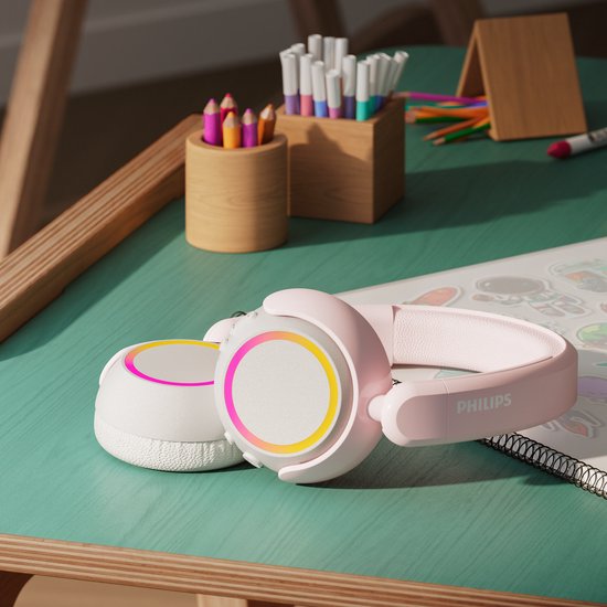 Roze en witte Philips koptelefoon met verlichte oorschelpen, op een bureau met tekenmateriaal.
