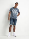 Jonge man met een blauw T-shirt met tekstprint en spijkerbroek shorts. Hij draagt witte hoge sneakers.
