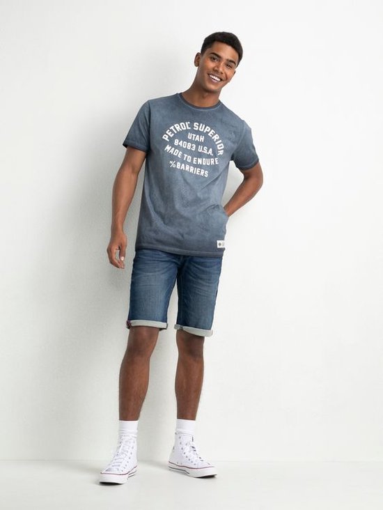 Jonge man met een blauw T-shirt met tekstprint en spijkerbroek shorts. Hij draagt witte hoge sneakers.