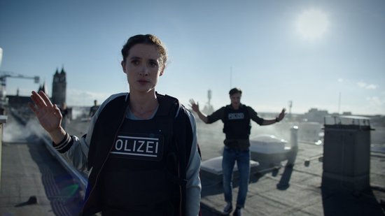 Vrouw met "POLIZEI" vest en man met "POLIZEI" vest op dak, met stad op achtergrond.