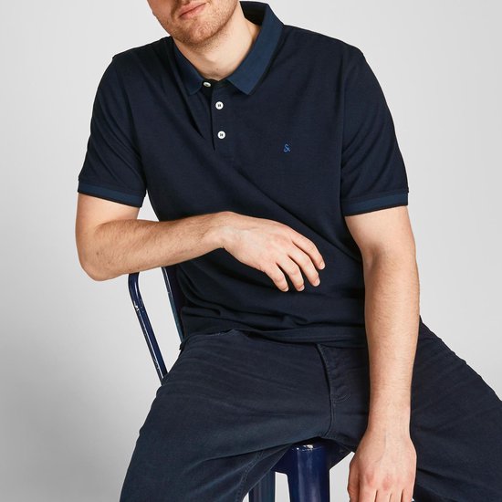 Man zit, draagt een donkerblauwe poloshirt met korte mouwen, donkerblauwe kraag, donkere broek.