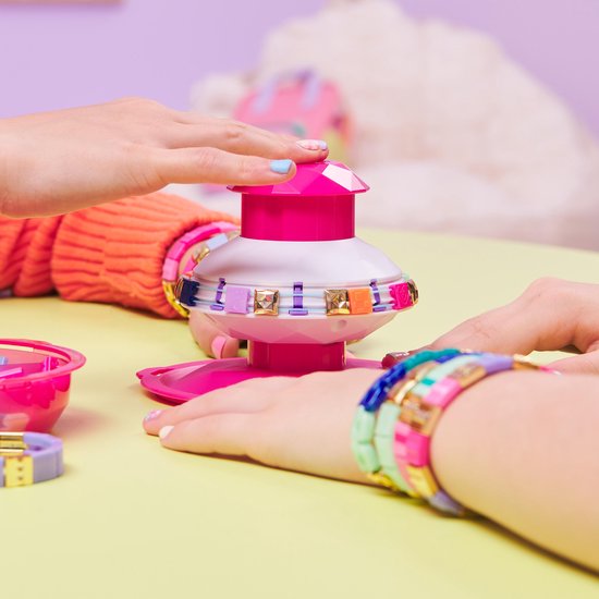 Roze en witte armbandenmaker met gekleurde parels, twee handen met zelfgemaakte armbanden. Zichtbaar zijn de tekst 'Pop Style' en het logo.