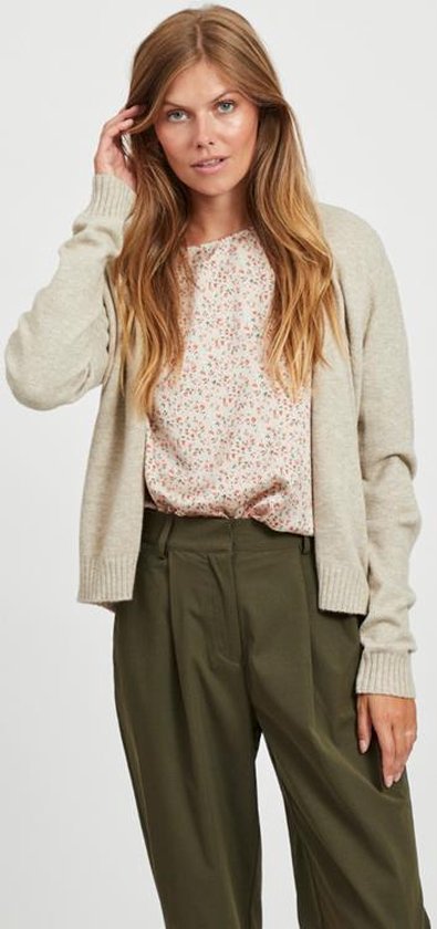 Model draagt een beige gebreid vest met een bloemenprint top en olijfgroene broek.