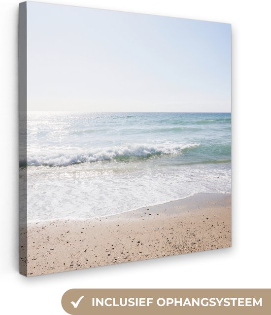 Een canvasafdruk van een zonnig strand met turquoise golven en zand, met de tekst 'Inclusief ophangsysteem'.