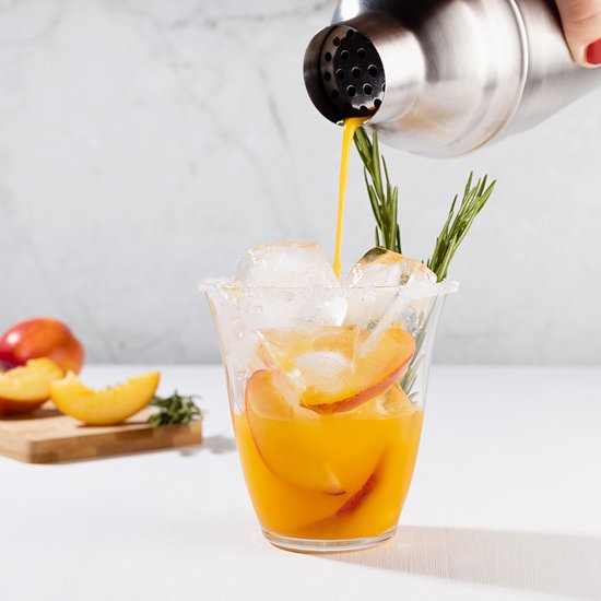 Buiten- en binnenrand van zout op een glas met ijsblokjes, perzikplakjes en takjes rozemarijn. Een cocktail wordt erin geschonken.