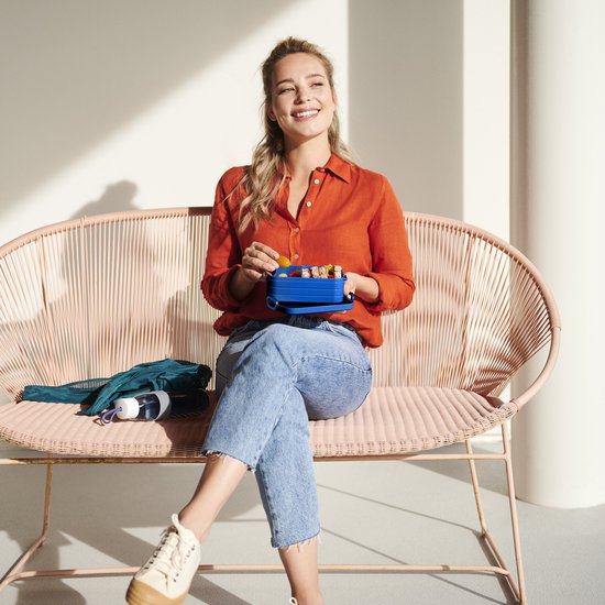 Femme en chemise orange et jean, assise, tenant une boîte à lunch bleue avec des amuse-gueules.