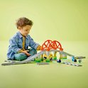 Jongetje zit op de grond te spelen met een LEGO Duplo treinbaan met een brug, overweg en twee poppetjes.