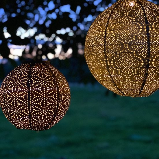 Twee verlichte gouden lampionnen met perforaties, hangend in de schemering tussen boomtakken.